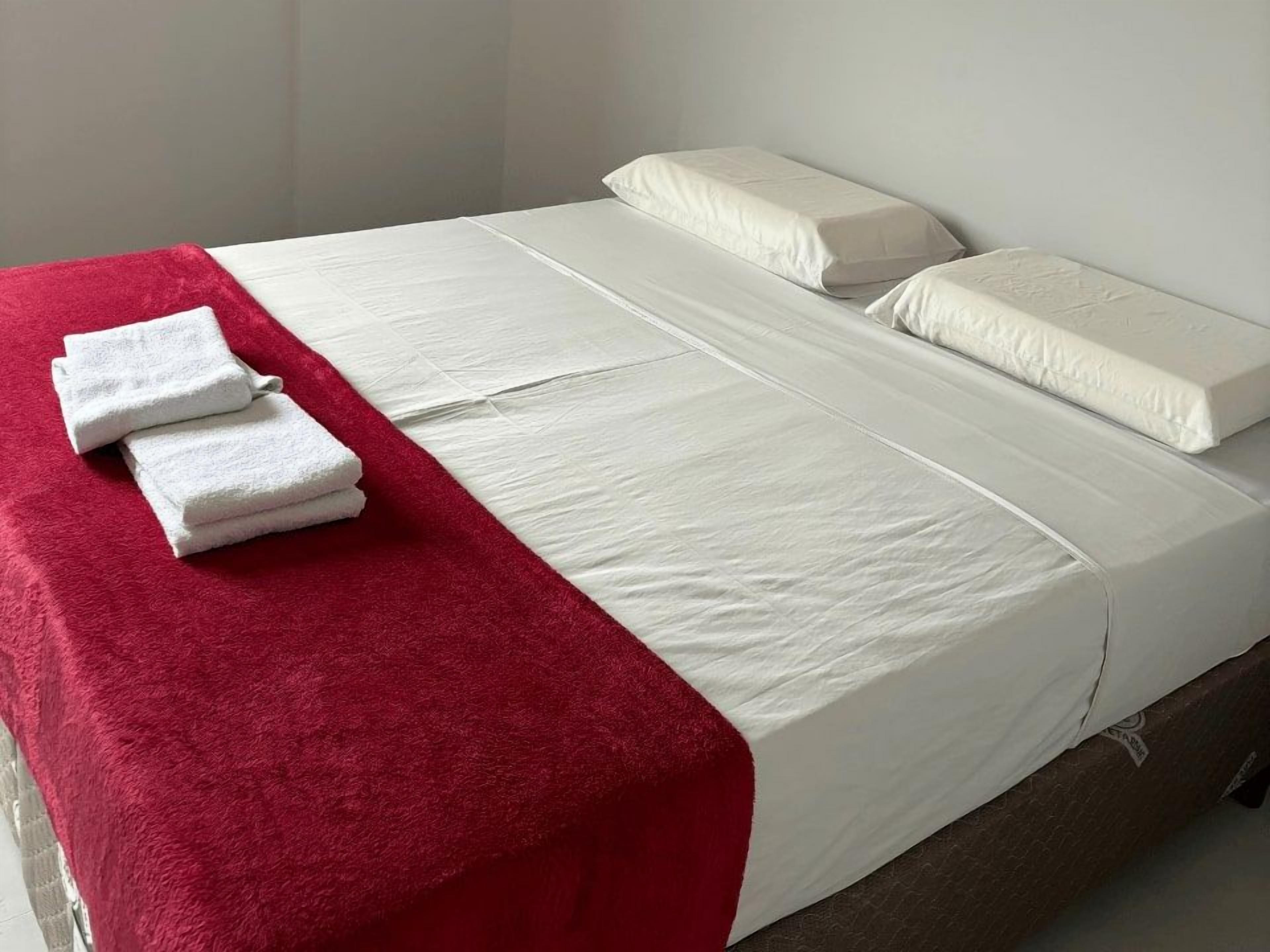 Una cama individual con ropa de cama blanca, una manta roja y dos toallas blancas a un lado.