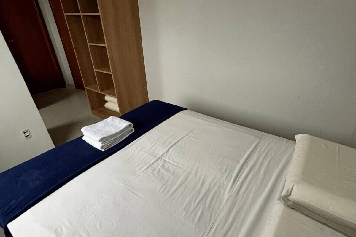 A neatly made bed with white linens and a blue bedspread, a wooden wardrobe, and a small stack of folded towels.