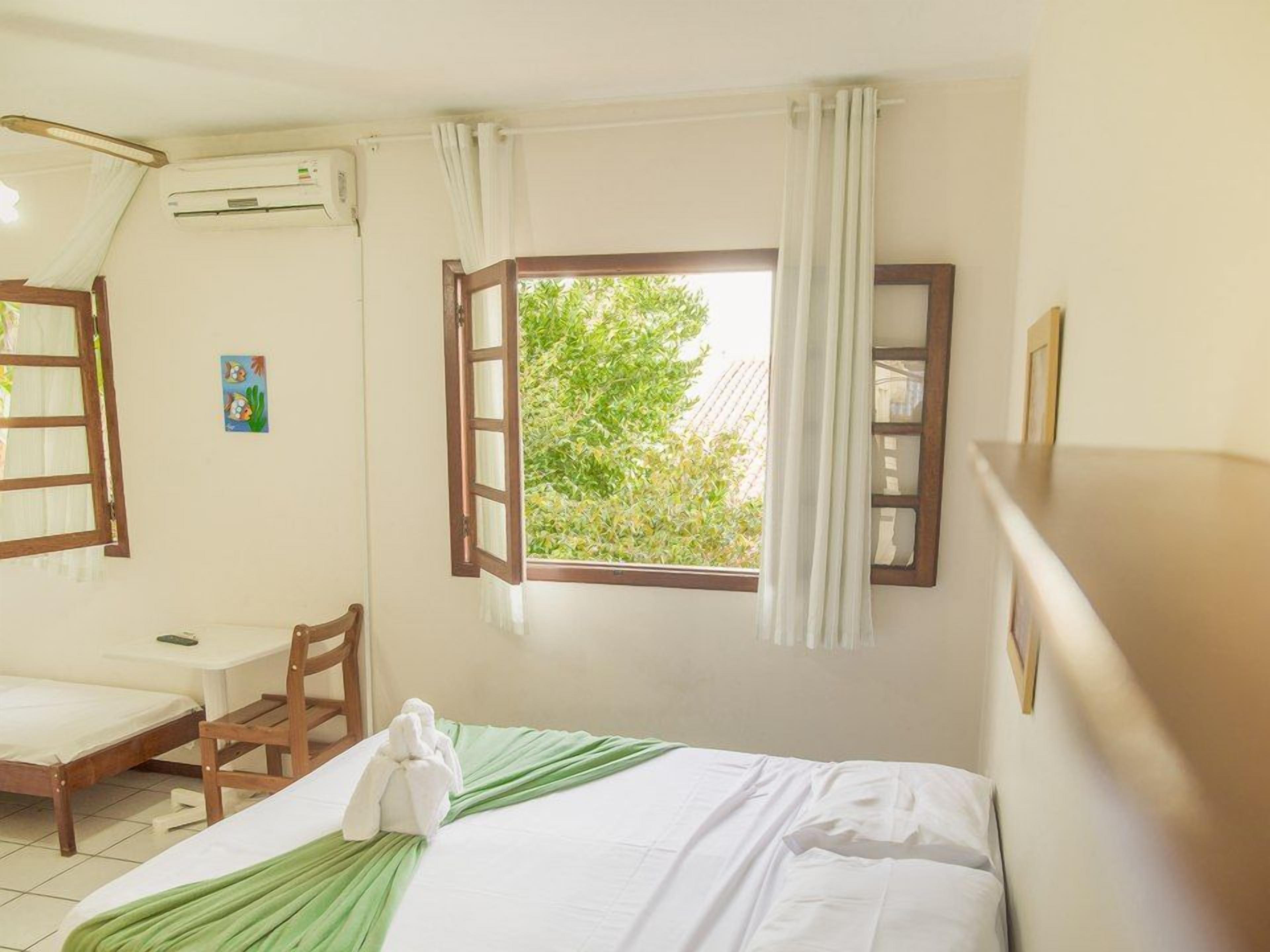 Una habitación con cama, escritorio y silla. Se ve un ventanal con cortinas, un equipo de aire acondicionado y un cuadro pequeño en la pared.