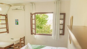 Een kamer met een bed, een bureau en een stoel. Er is een raam met gordijnen, een airconditioning en een klein schilderij aan de muur.