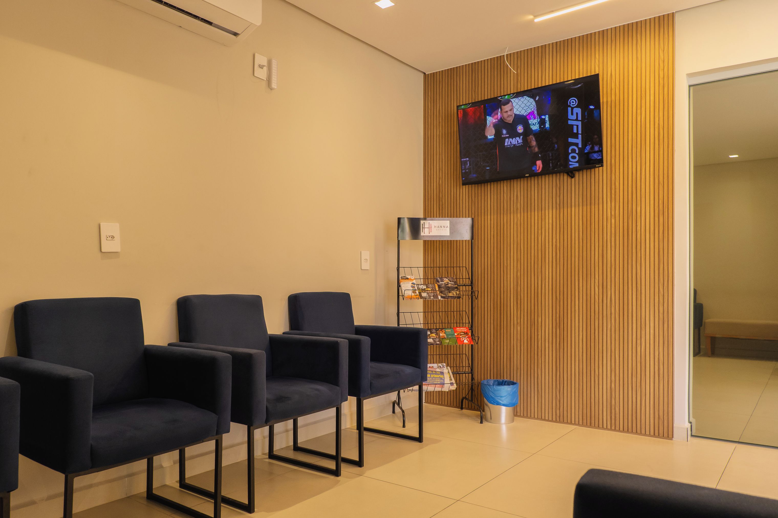 Un área de recepción con un televisor de pantalla plana montado en la pared, paneles de madera y varias sillas azul oscuro dispuestas para esperar.