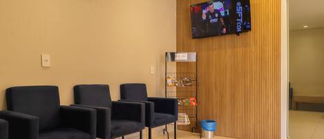 A reception area with a flat-screen TV mounted on the wall, a wooden panel wall, and several dark blue chairs arranged for waiting.