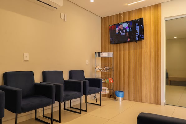 A reception area with a flat-screen TV mounted on the wall, a wooden panel wall, and several dark blue chairs arranged for waiting. - Hanna Amsterdam Hotel & Eventos (Florianópolis)