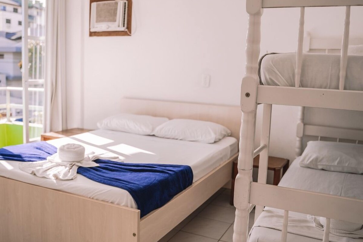 Habitación con una cama individual y literas de madera, ropa de cama blanca, una manta azul y una ventana con vistas a edificios.