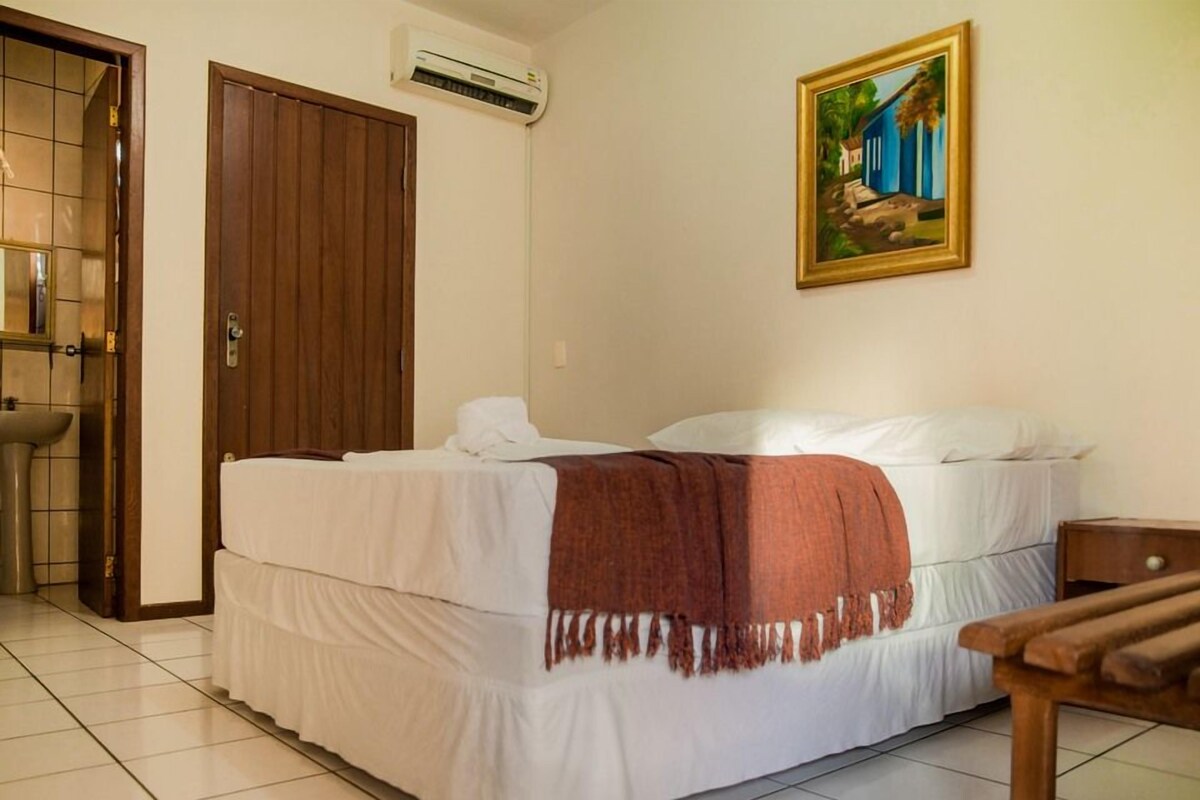 A hotel room with a bed, a painting on the wall, a wooden door, and a bathroom with a sink.