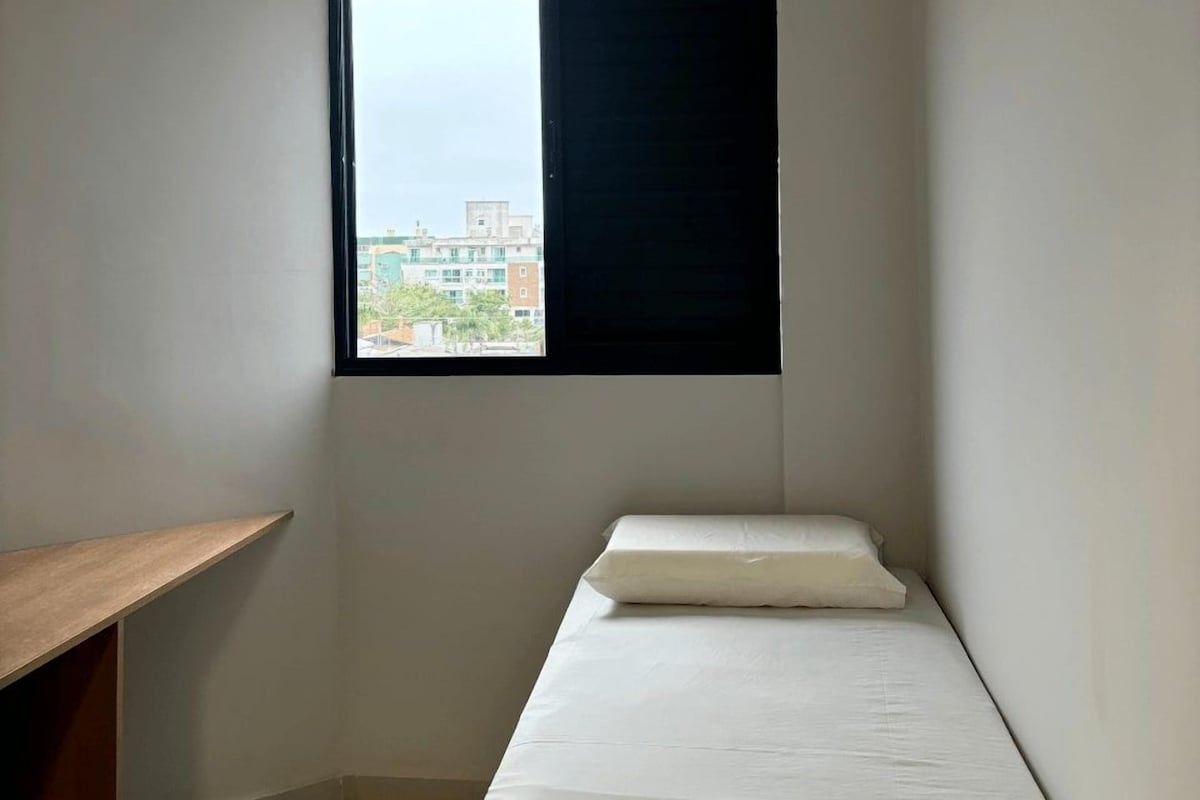 A small, single-bed room with a window, a wooden desk, and a view of buildings outside.