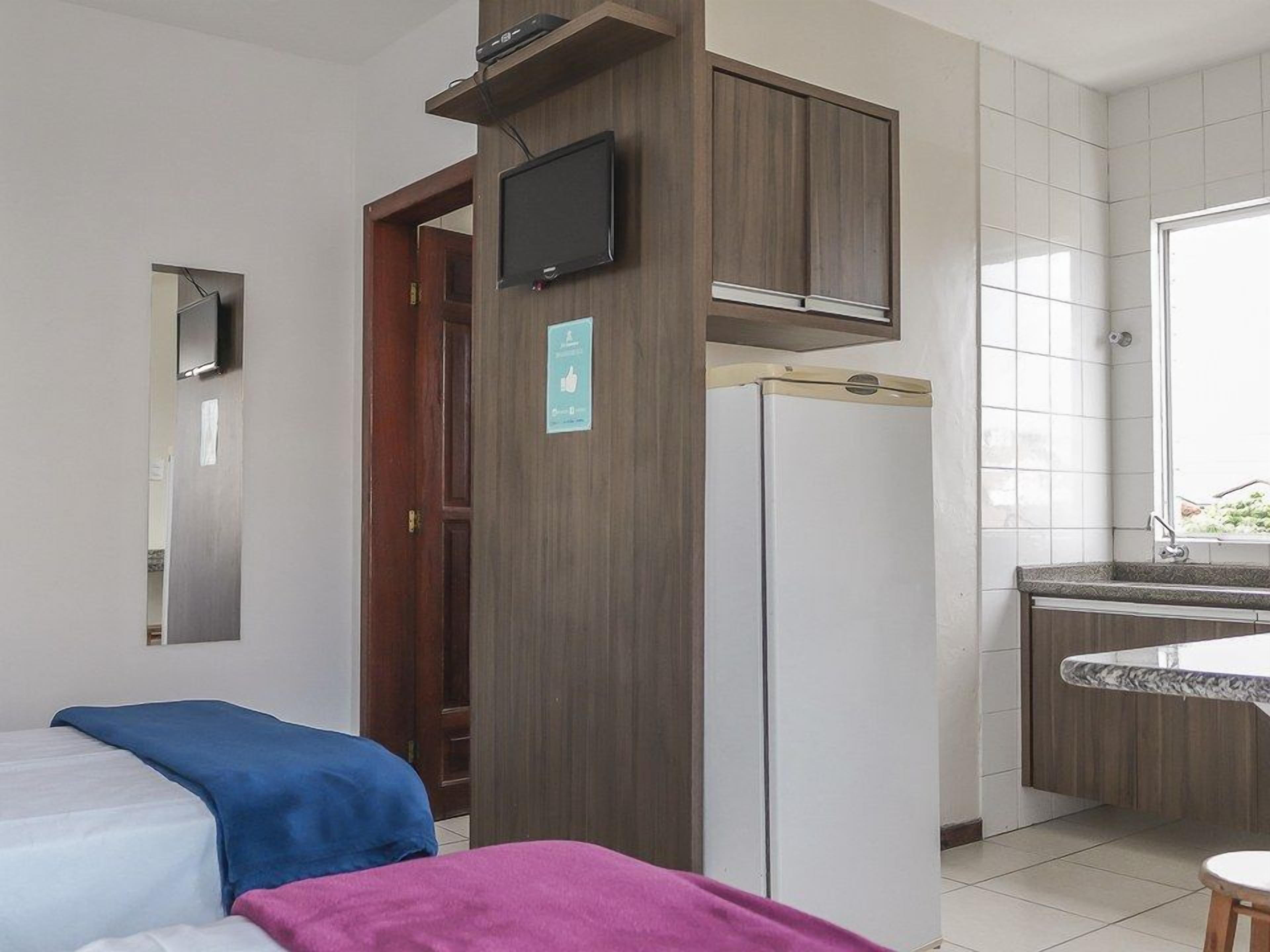 A hotel room with a bed, a small kitchenette, a TV mounted on the wall, and a window.