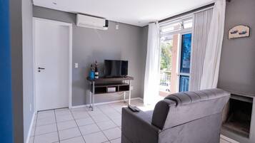 A modern living room with a grey sofa, a flat-screen TV on a wooden stand, and a balcony with a view.