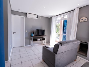 A modern living room with a grey sofa, a flat-screen TV on a wooden stand, and a balcony with a view. - Hanna Amsterdam Hotel & Eventos (Florianópolis)