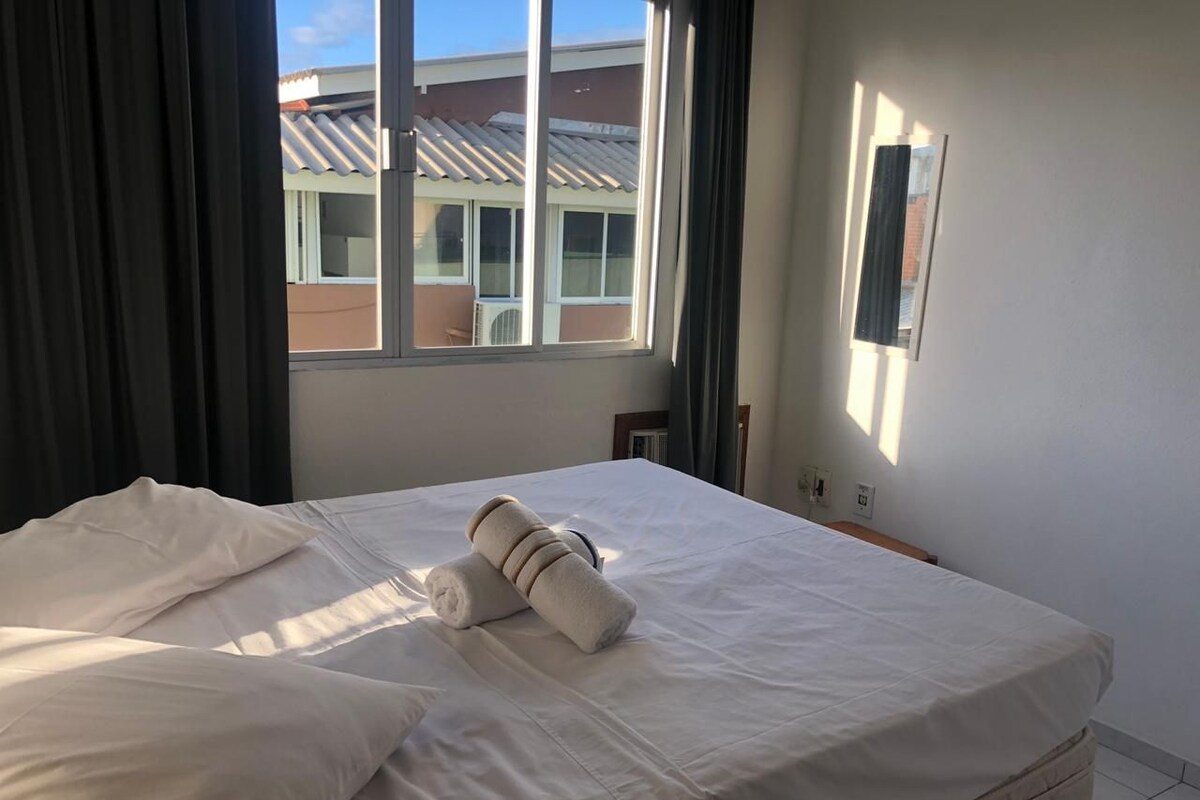 A neatly made bed with white linens and a rolled towel, a window with curtains, and a wall-mounted light switch.