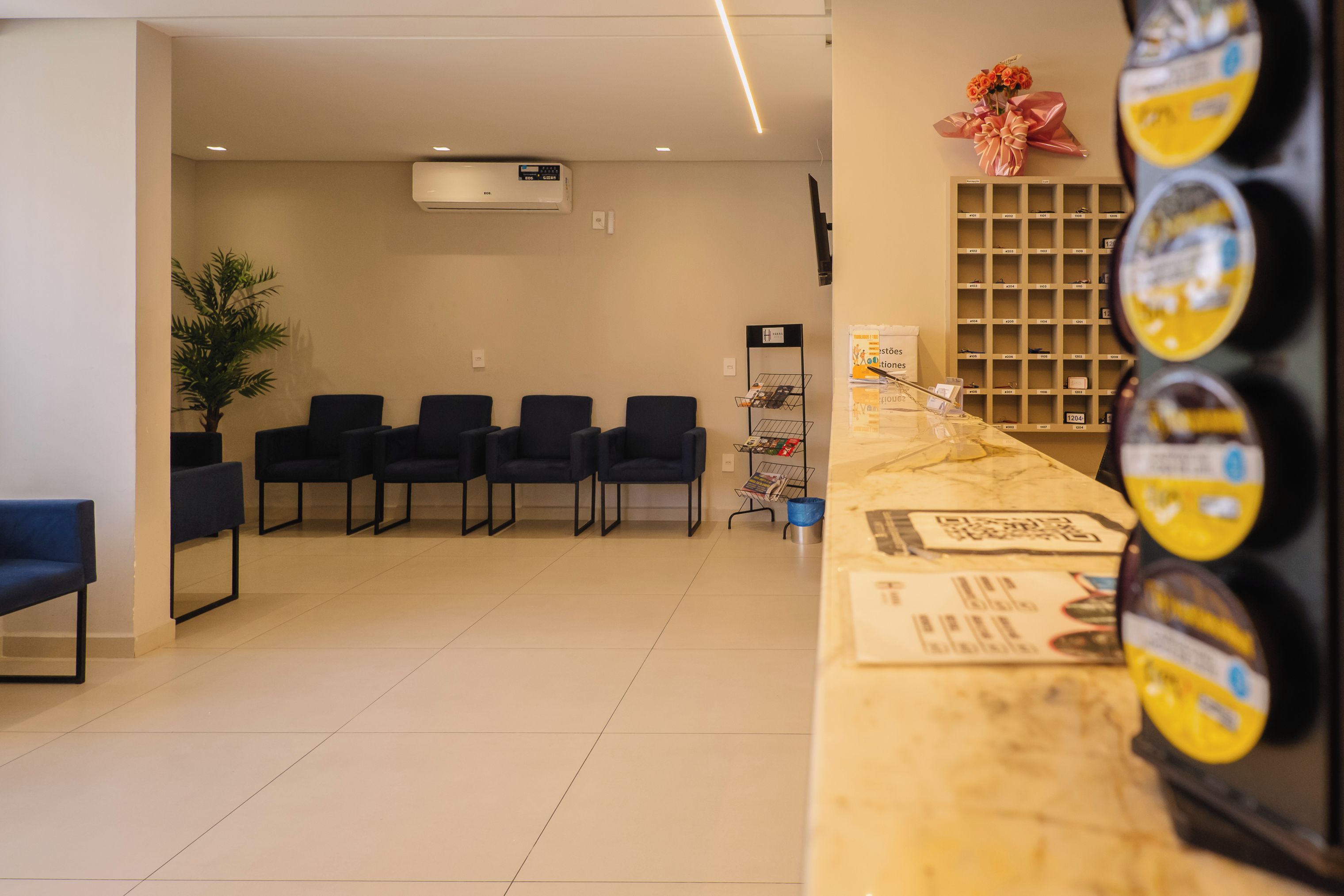 A reception area with a counter, chairs, a potted plant, and a wall-mounted shelf with items.