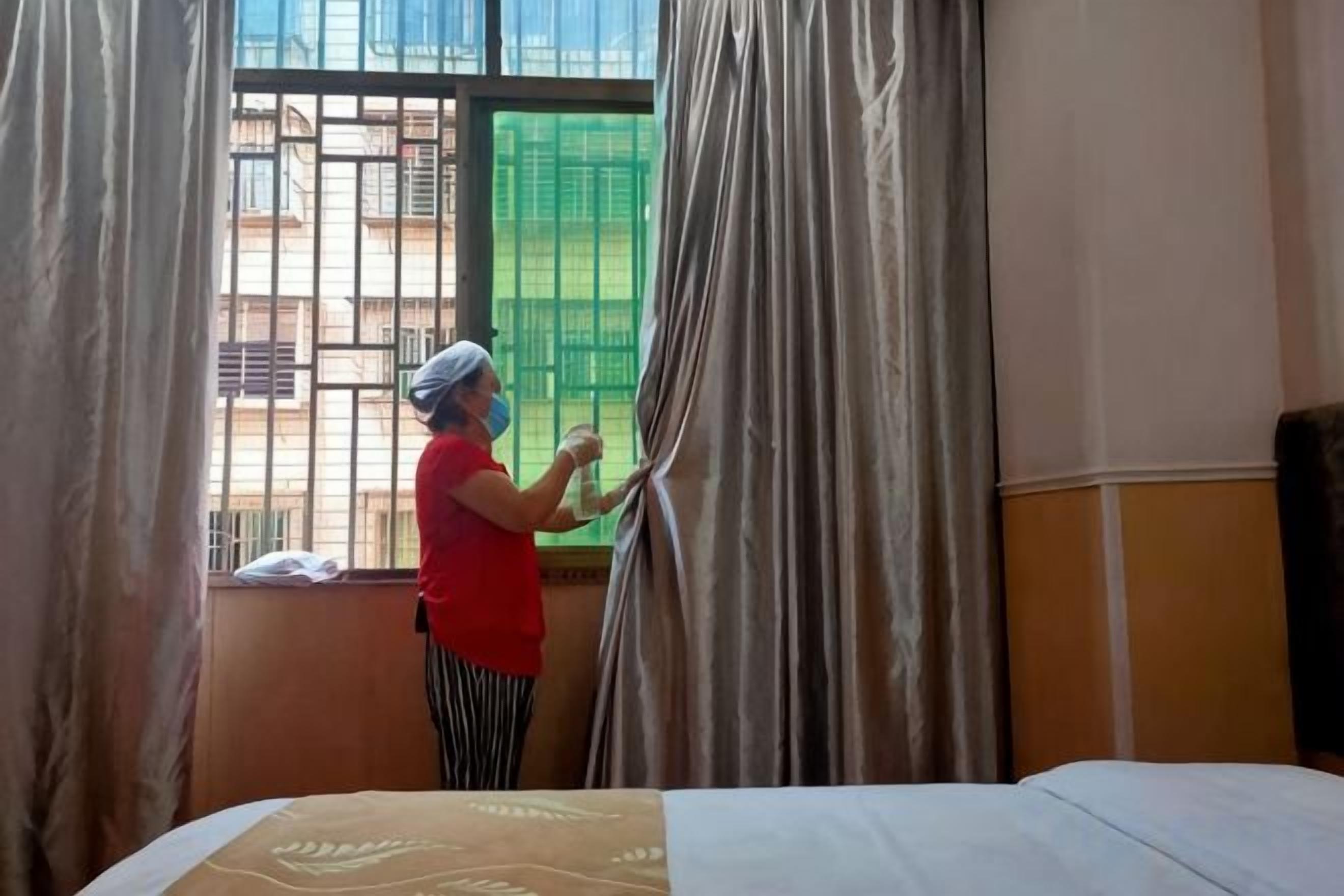 A person adjusting curtains in a room with a bed and a window.