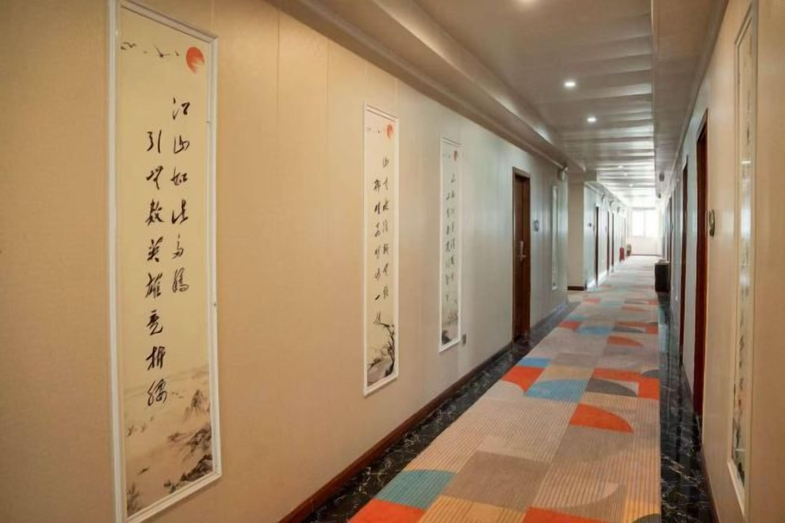 A hotel corridor with a patterned carpet, framed calligraphy on the walls, and doors leading to rooms.