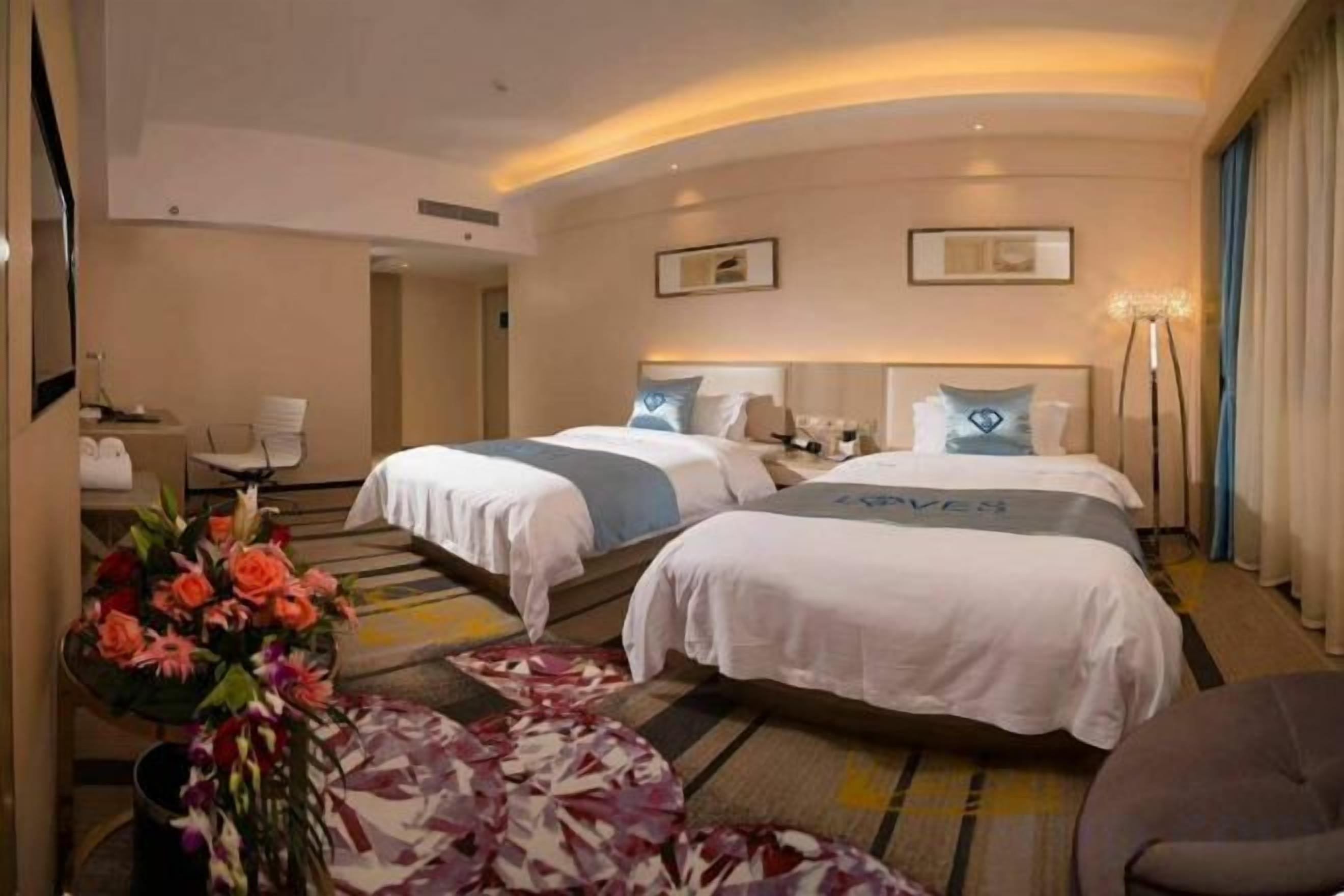 A hotel room with two beds, a desk, a chair, and a vase of flowers.
