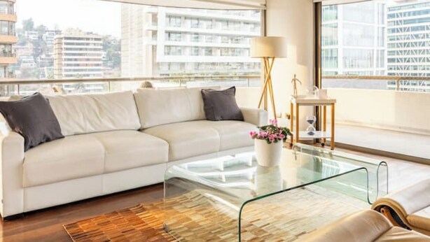 Ein modernes Wohnzimmer mit einem weißen Sofa, einem Glastisch und Blick auf eine Stadtlandschaft durch große Fenster.