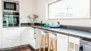 Eine moderne Küche mit Einbauofen, Mikrowelle und Kühlschrank. Weiße Barhocker und ein Fenster mit Blick auf einen Baukran.