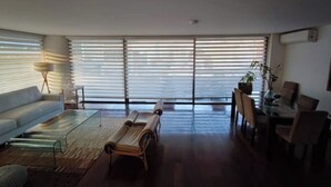 Ein modernes Wohnzimmer mit einer Couch, einem Couchtisch und einer Bank. Es gibt große Fenster mit Jalousien, einen Essbereich mit Stühlen und eine Pflanze.