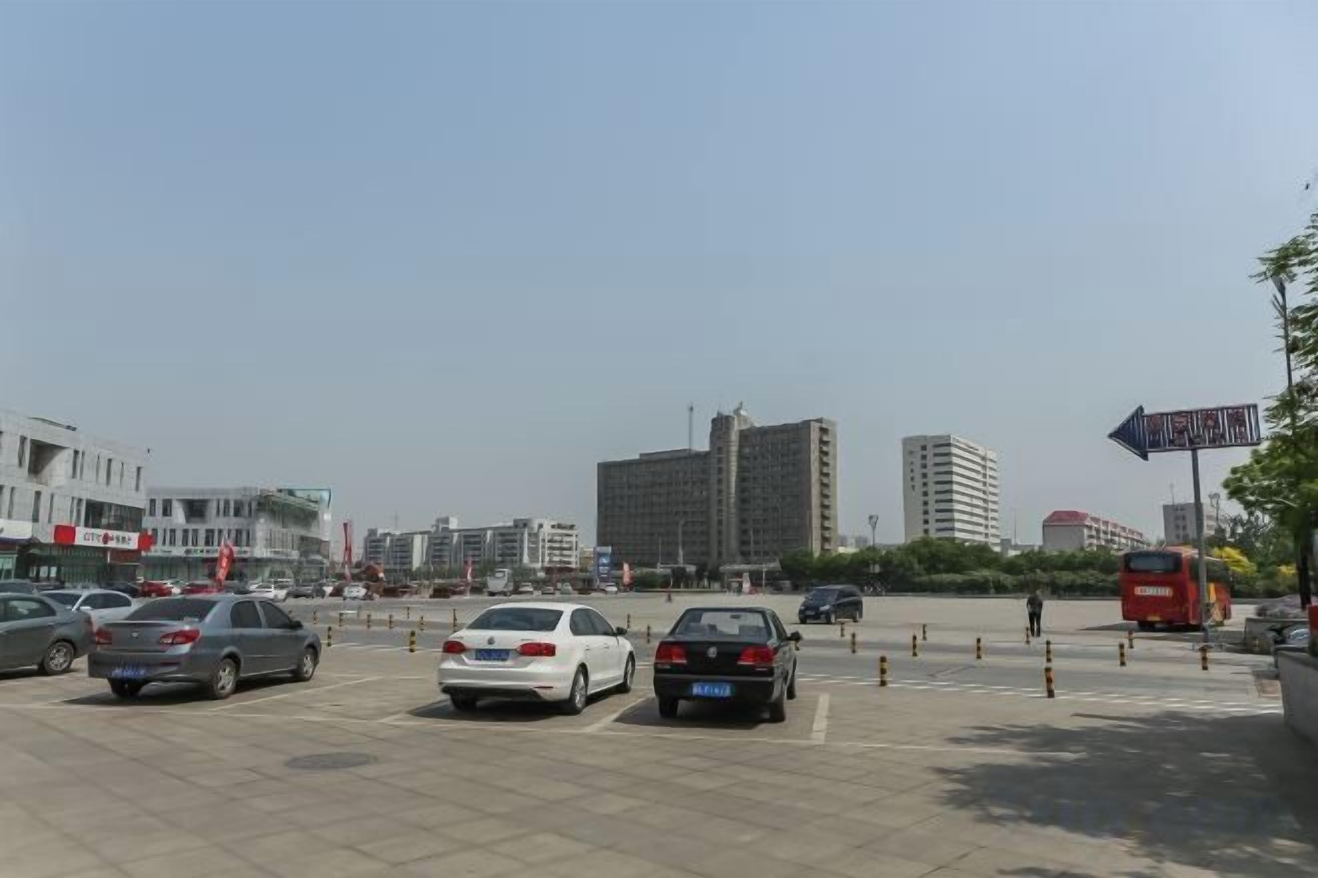 A parking lot with multiple cars and a red bus, surrounded by buildings and a clear sky.
