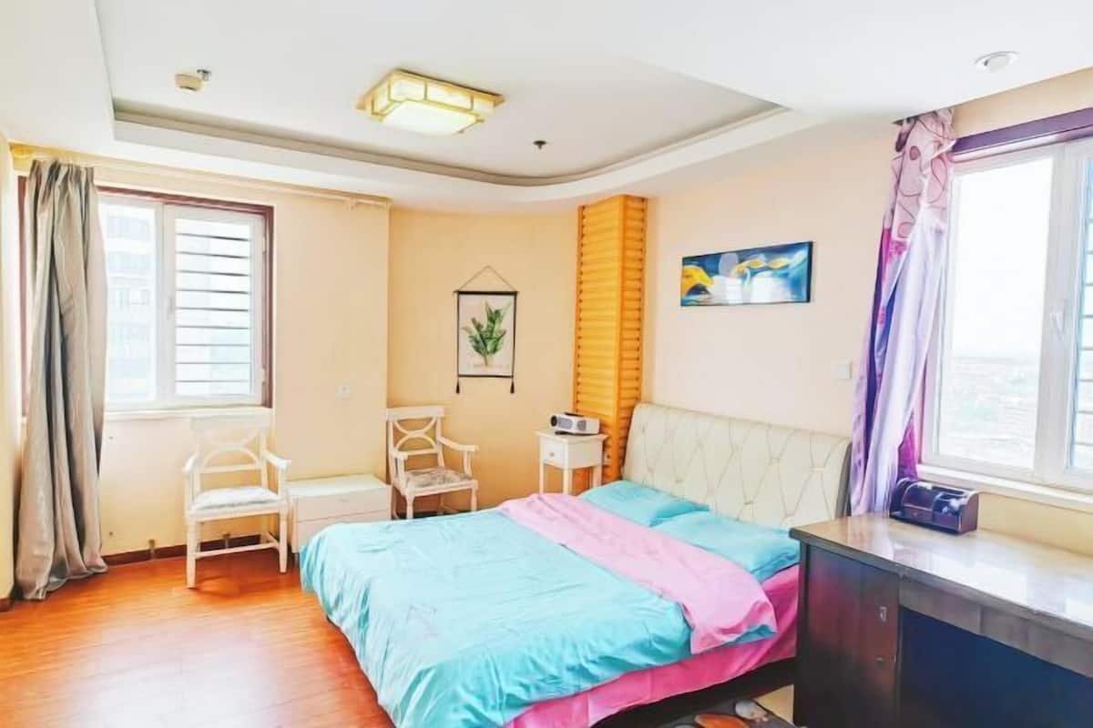 Een slaapkamer met een bed, een bureau, een stoel, een raam met gordijnen en een aan de muur hangend schilderij.