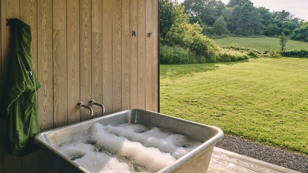 A wooden cabin with a metal sink filled with bubbles, a green towel hanging, and a view of a green field and trees.