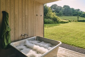 A wooden cabin with a metal sink filled with bubbles, a green towel hanging, and a view of a green field and trees.