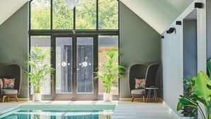 Un espace piscine intérieur moderne, doté d’un haut plafond et de grandes fenêtres, d’une douche intégrée et de coins salon agrémentés de plantes.