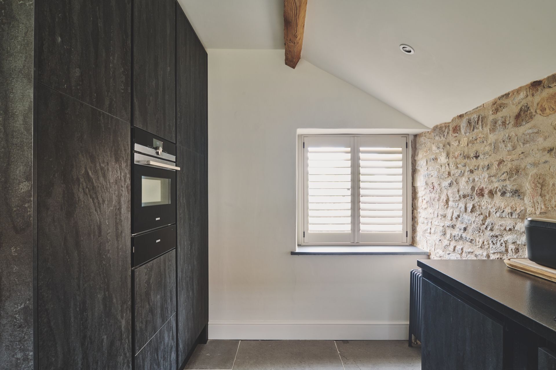 Une cuisine avec des meubles de rangement aux couleurs sombres, un four encastré, une fenêtre avec des stores et un mur en pierre.