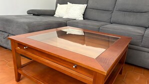 Una mesita de madera con tapa de cristal, ubicada frente a un sofá gris con un cojín blanco.