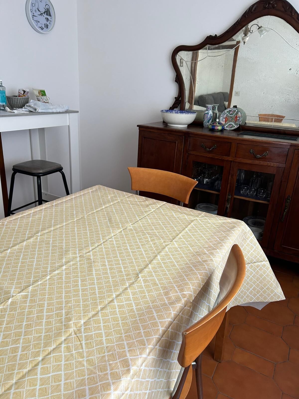 Un área de comedor con una mesa cubierta por un mantel estampado, dos sillas, un aparador de madera con puertas de cristal y un reloj de pared.