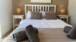 A neatly made bed with a white headboard, two bedside tables with lamps, and a large abstract painting on the wall. - Almirida Paradise Apartments (Chania)