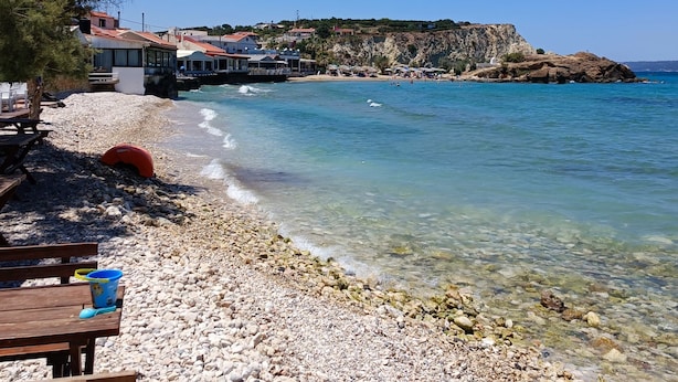 A pebble beach with clear water, a wooden bench, and a view of buildings and cliffs in the background. - Almirida Paradise Apartments (Chania)