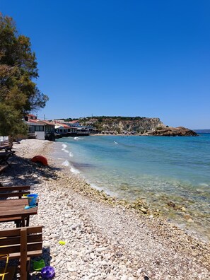 A pebble beach with clear water, a wooden bench, and a view of buildings and cliffs in the background. - Almirida Paradise Apartments (Chania)