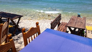 A beach with clear water, wooden tables and chairs, and a distant mountain. - Almirida Paradise Apartments (Chania)