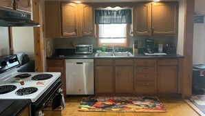 A compact kitchen with wooden cabinets, a stove, a dishwasher, and a window with curtains. - Rustic cottage by the mighty Elk! (Girard)