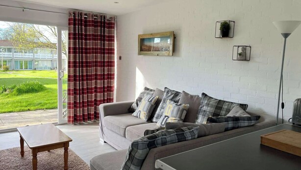 A modern living room with a grey sofa, plaid cushions, a wooden coffee table, and a view of a green garden through a glass door.