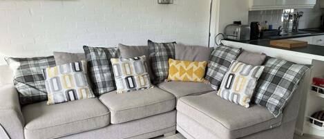 A grey sectional sofa with multiple cushions in various patterns, a white brick wall, a small framed painting, and a kitchen area in the background.