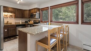 A kitchen with granite countertops, wooden cabinets, a built-in island with bar stools, a microwave, and a view of trees through large windows. - Spacious Townhome on Shuttle Route, Just Minutes to Vail Resort! (Vail)