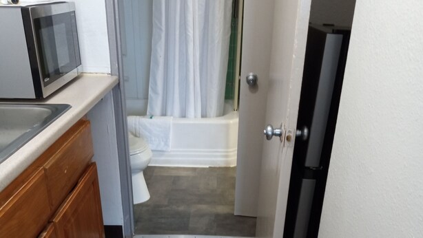 Un couloir étroit mène à une salle de bain équipée de toilettes, d’une baignoire et d’un rideau de douche. On peut apercevoir un four à micro-ondes sur le côté gauche.