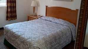 Un lit bien fait, recouvert d’une couvre-lit à motifs, une table de chevet avec une lampe, un tableau représentant une scène de plage et une fenêtre avec des rideaux.