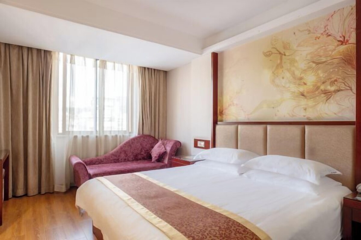 Een hotelkamer met een groot bed, een kleinere zithoek, een schilderij aan de wand en een raam met gordijnen.