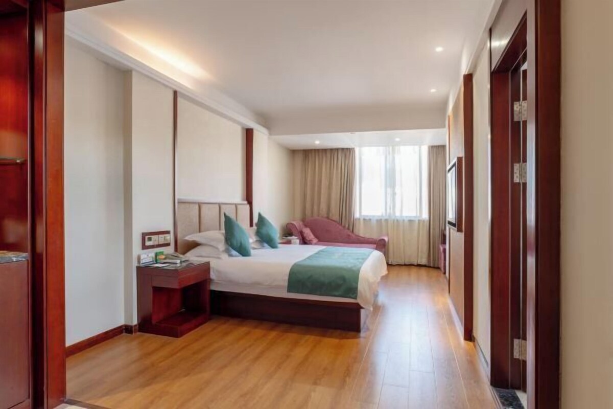 Een hotelkamer met een houten vloer, een bed met groen en witte beddengoed, een nachtkastje en een raam met gordijnen.