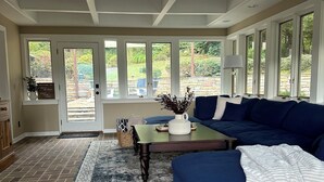 Een ruime woonkamer met een grote blauwe bank, een salontafel en een kamerbreed tapijt. Er is een deur die leidt naar een buitenruimte met uitzicht op groen.