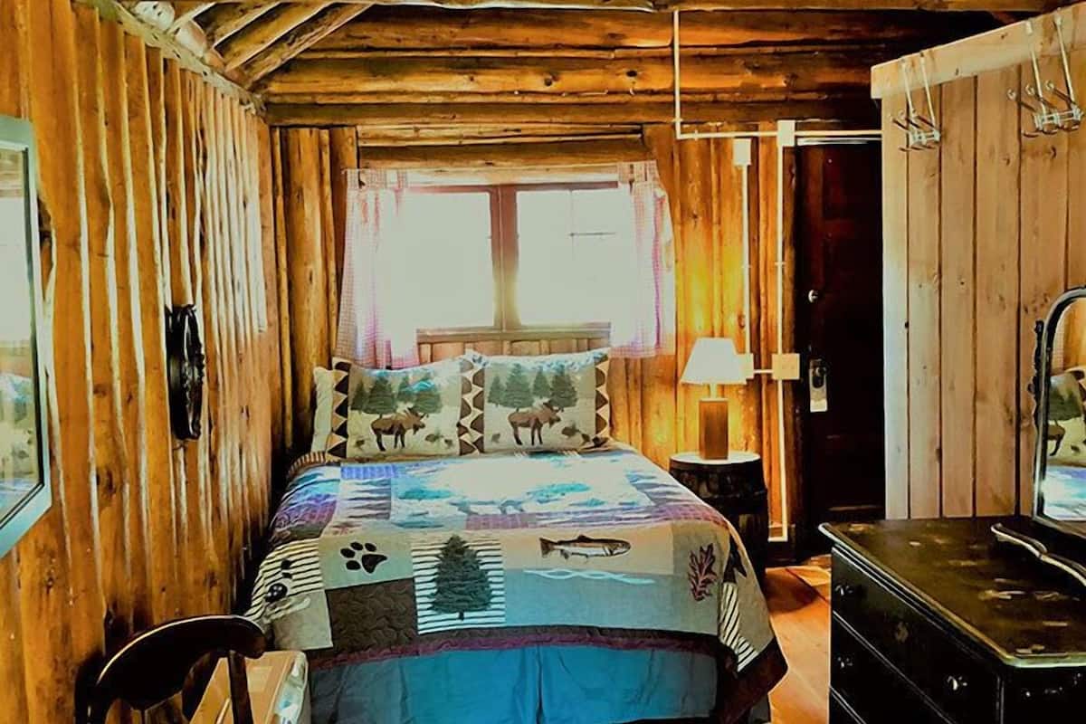 A wooden cabin bedroom with a bed, a dresser, a chair, and a window with curtains.