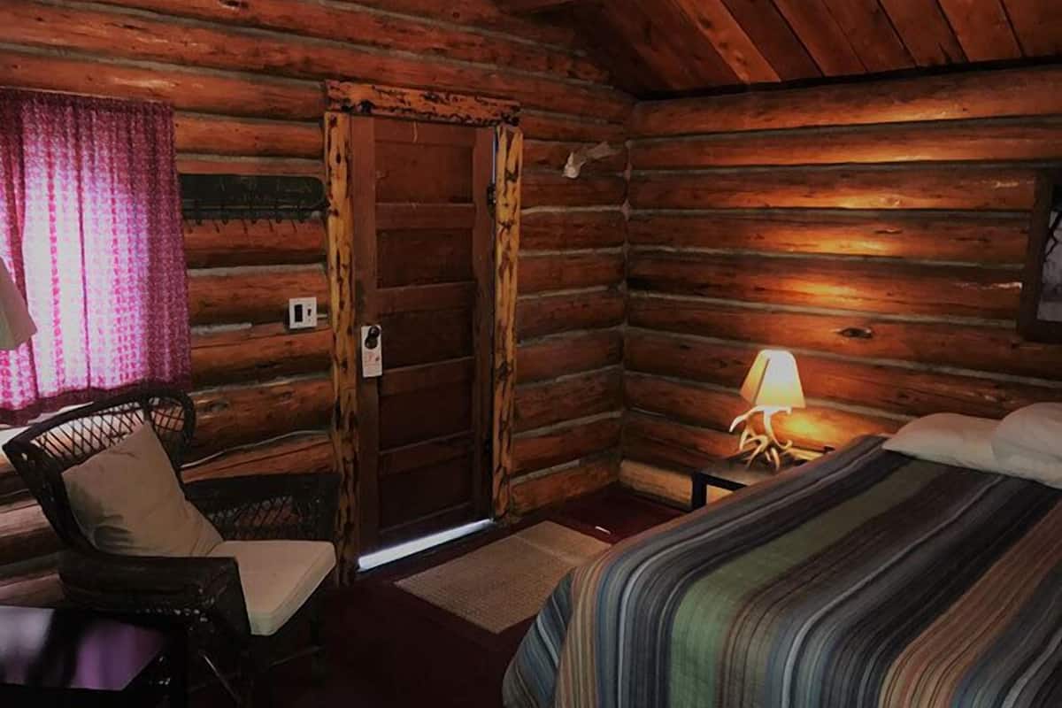 A log cabin room with a bed, a chair, a table, and a door.