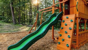 Une structure de jeux en bois avec un toboggan vert et un mur d’escalade, située dans une zone boisée.