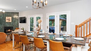 Une salle à manger moderne avec une longue table dressée pour six personnes, entourée de chaises, d’une cheminée et offrant une vue sur l’extérieur.