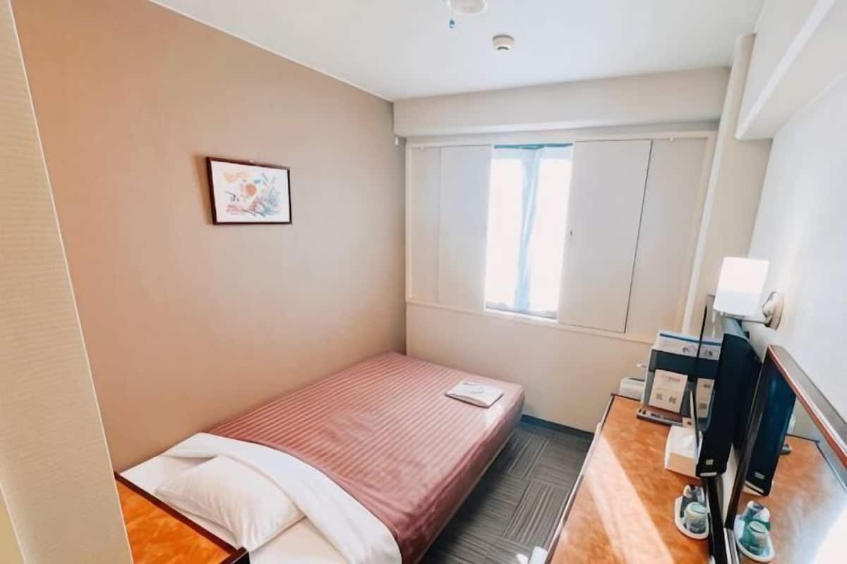 A small, single-bed room with a wooden floor, a bedside table with a lamp, a window with curtains, and a framed picture on the wall.