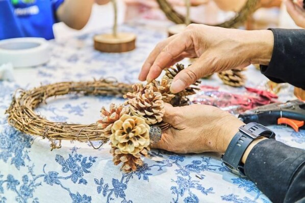 Hands arranging a pine cone wreath on a table with a floral tablecloth.