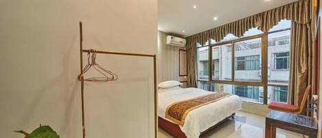 A hotel room with a bed, a wooden hanger rack, a potted plant, and a large window with curtains.