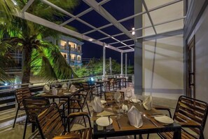 Un espace de restauration en plein air avec des tables prévues pour deux personnes, entouré de chaises en bois et séparé par une cloison en verre.